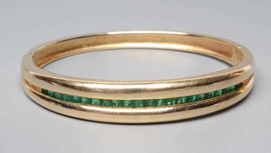 AN EMERALD STIFF HINGED BANGLE, the upper section channel set with twenty two graduated calibre