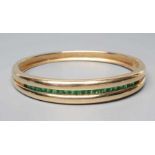 AN EMERALD STIFF HINGED BANGLE, the upper section channel set with twenty two graduated calibre