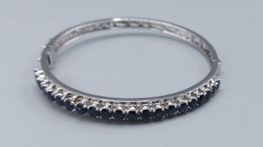 A white metal bangle set with a row of twenty one sapphire, claw set, 12.9gms, 6.5cms diameter
