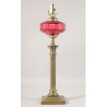 A BRASS CORINTHIAN COLUMN OIL LAMP with cranberry glass reservoir, (converted to electricity). 22ins
