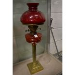 A brass and cranberry coloured glass oil lamp with a shade.