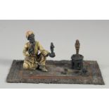 A COLD PAINTED BRONZE GROUP, the amulet vendor sitting on a rug. 7ins long.