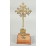 A BRASS CROSS on a wooden stand, with gemstones. 11.5ins high