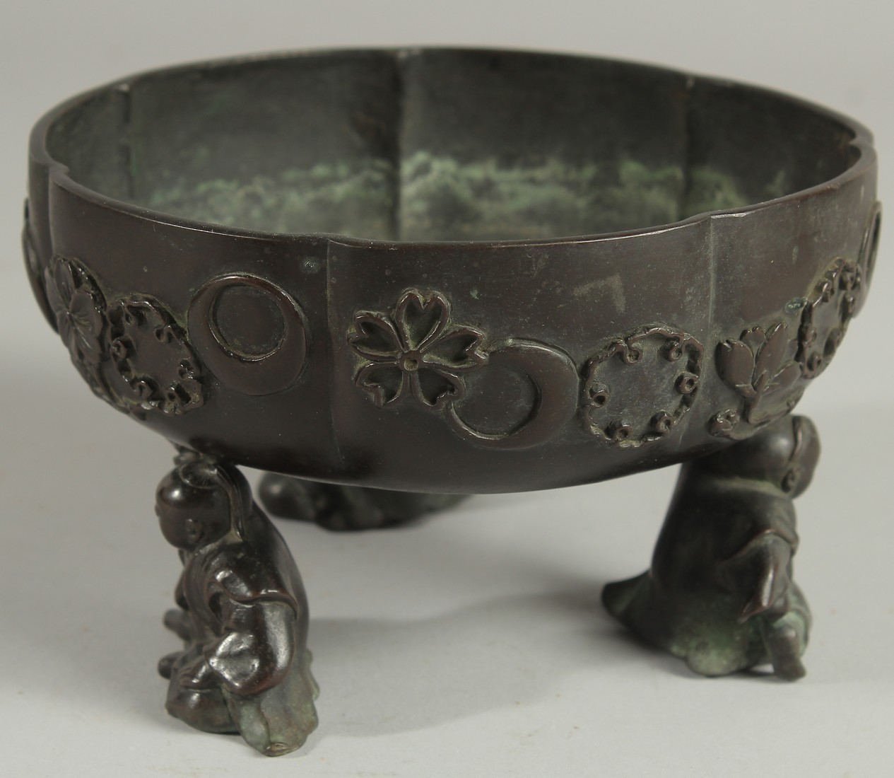 A JAPANESE BRONZE PETAL FORM BOWL, raised on three mounted figural feet, the bowl with relief mon