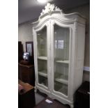 A French carved and white painted armoire with scroll crest over two arch top glazed doors, on