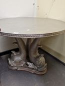 A CONTEMPORARY SILVERED CIRCULAR TABLE SUPPORTED ON A THREE DOLPHIN COLUMN AND TRIPARTITE FOOT.