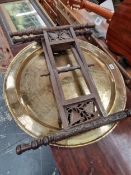 AN EASTERN BRASS CIRCULAR DISH FORMING A TABLE TOP WITH ITS FOLDING CARVED WOOD STAND