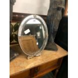 AN EASEL BACKED OVAL MIRROR, THE FRAME CUT WITH A BAND OF ROUNDELS AND FIXED WITH FLOWER HEADS