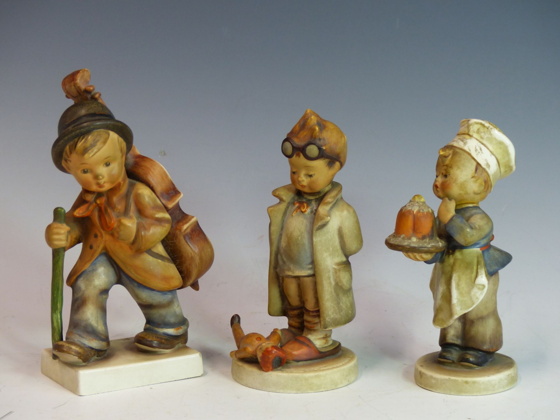 THREE M HUMMELL FIGURINES, A YOUNG CHEF WITH CAKE, A BOY WITH DOLL AT HIS FEET & A YOUNG MUSICIAN