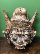 A BURMESE CARVED AND PAINTED MASK OF A MYTHICAL BEAST WEARING A WINGED HELMET ABOVE ITS PROTRUDING