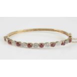 A 9ct yellow gold, ruby and diamond hinged oval hollow bangle, featuring eight round faceted