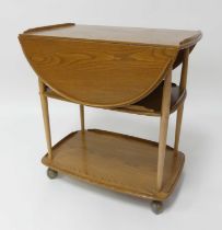 An Ercol blond elm dropflap three-tier tea trolley, each tier with raised border upon roller