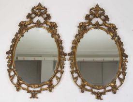 A pair of Rococo style gilt composition framed and bevelled oval wall mirrors, the swept frames with