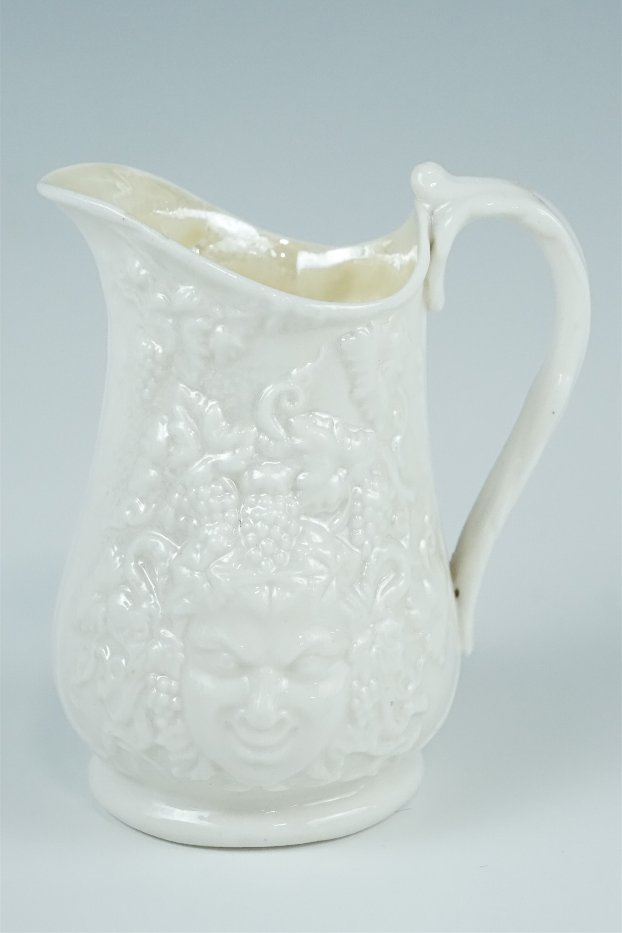 A Belleek cream jug, relief decorated with Bacchus masks and grape vines, (free from damage and