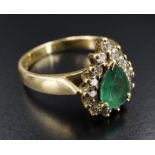 An emerald and diamond ring, having a 7 x 5 mm pendeloque cut emerald, surrounded by 15 brilliant