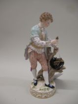 Meissen figure of a young man wearing a pale blue tunic gathering sticks in his hat, standing beside