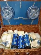 A wicker picnic basket, containing plates, cups, and flasks.