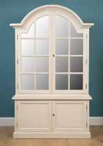 A cream painted OKA display cabinet with an arched top above glass panelled doors, supported by a