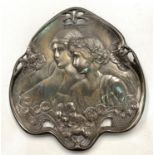 An early 20th century silvered metal plaque, spade shaped with relief decoration of a young couple