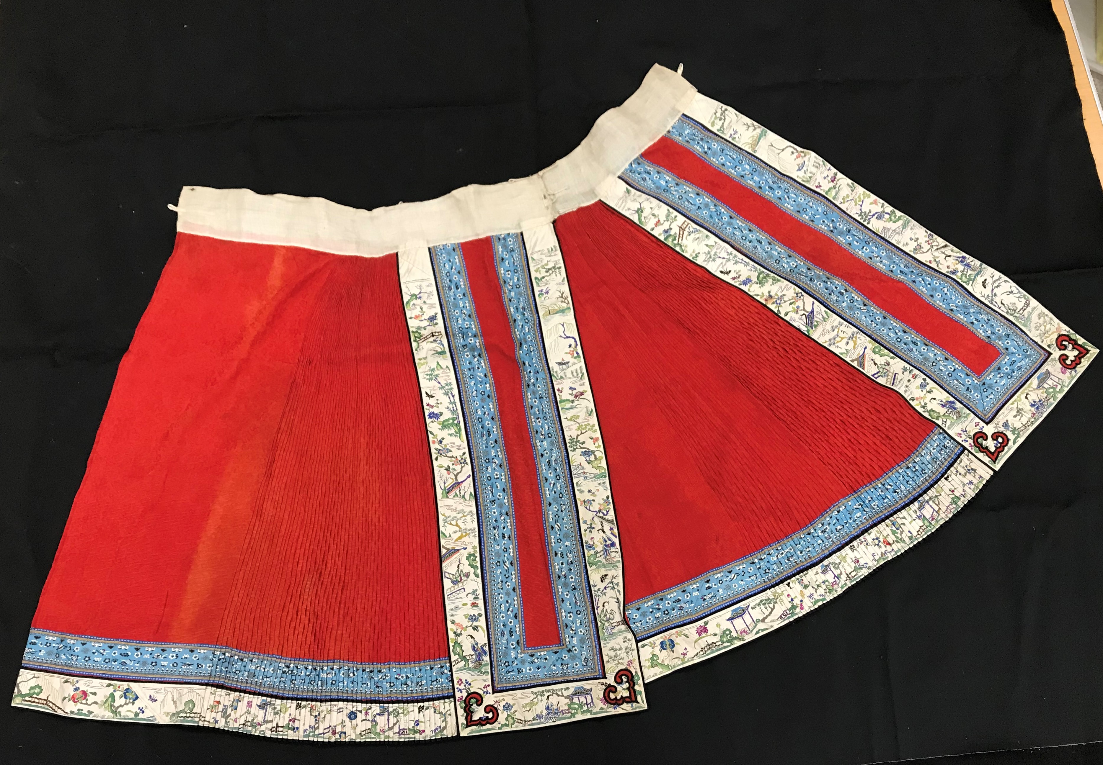 A Chinese red finely pleated silk skirt with blue and cream overlaid silk bands heavily embroidered