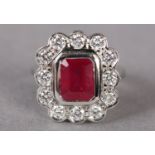 A RUBY AND DIAMOND CLUSTER RING IN PLATINUM, collet set to the centre with a step cut ruby within