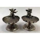 A pair of white metal shell salts (tests as silver), the bowls surmounted with cherub musicians,