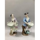A pair of German china figures in 18th century garb each carrying a basket and on bocage base,