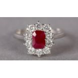 A RUBY AND DIAMOND CLUSTER RING in 9ct white gold, claw set to the centre with a step cut ruby