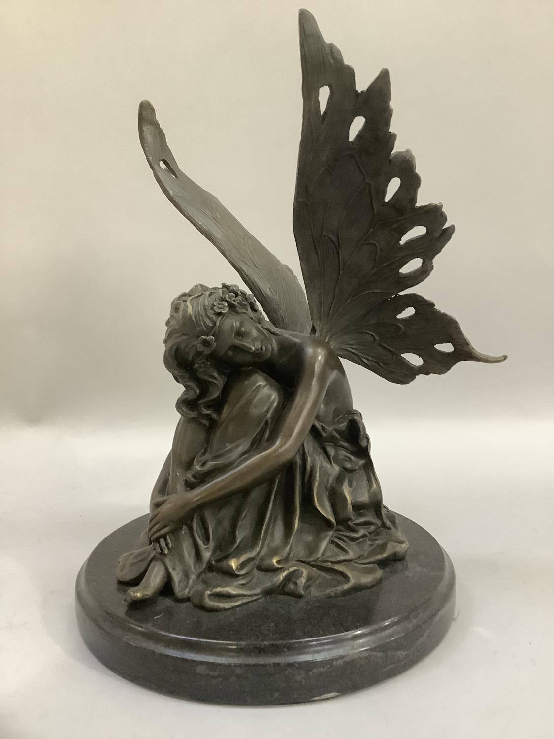A modern bronze-effect figure of a fairy, sitting resting her head on her knees and on a marble-type