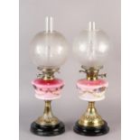 A near pair of Victorian brass oil lamps, the pink tinted reservoirs painted with mauve flower heads