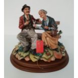 CAPODIMONTE FIGURINE depicting an old couple sat on a park bench, the gentleman smoking his pipe,