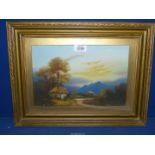 A gilt framed oil painting of a thatched cottage under tall trees with rugged mountains in the