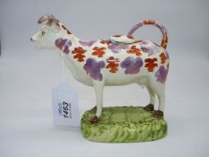 A circa 1830's pottery cow creamer with red and pink lustre patches, standing on green base,