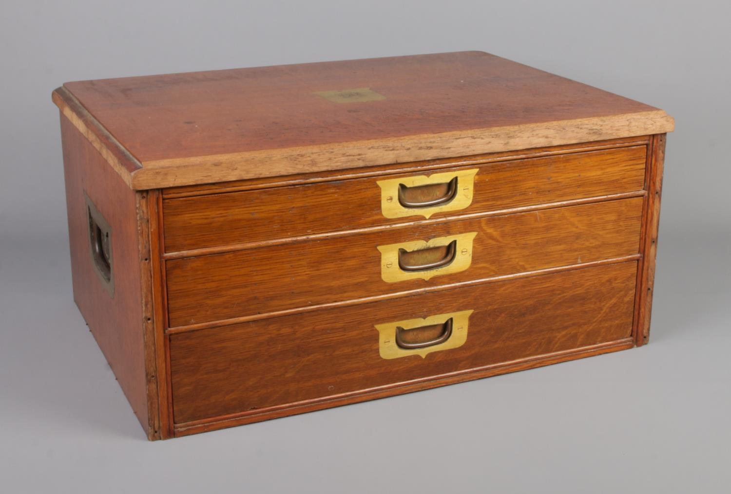 An oak tabletop three drawer display cabinet featuring campaign handles to front and sides.