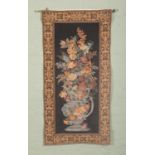 A brass mounted wall tapestry, depicting a floral display within an urn, with scrolled patterned