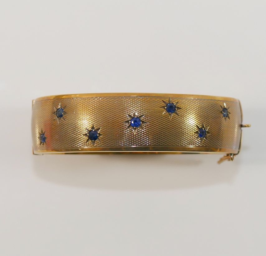 A 9 carat gold hollow hinged sapphire set bangle, with engine turned decoration, the seven round