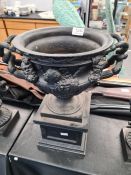 An old cast iron urn on square pedestal base, the body decorated heads, height 40cms