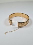 9ct yellow gold hinged bangle with floral engraved decoration, and safety chain, AF, 6cm diameter,