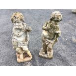 A pair of composition stone putti, the draped cherubs playing pipes standing by logs on square