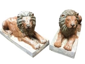 A pair of composition lions, the painted animals lying in prone positions on rectangular plinths. (