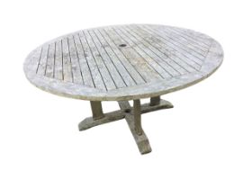 A circular teak garden table with slatted top on four rectangular supports joined by shaped sledge