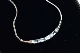 A 9CT GOLD, TOPAZ NECKLACE, a solid white gold crecent shaped bar, set with four emerald cut blue