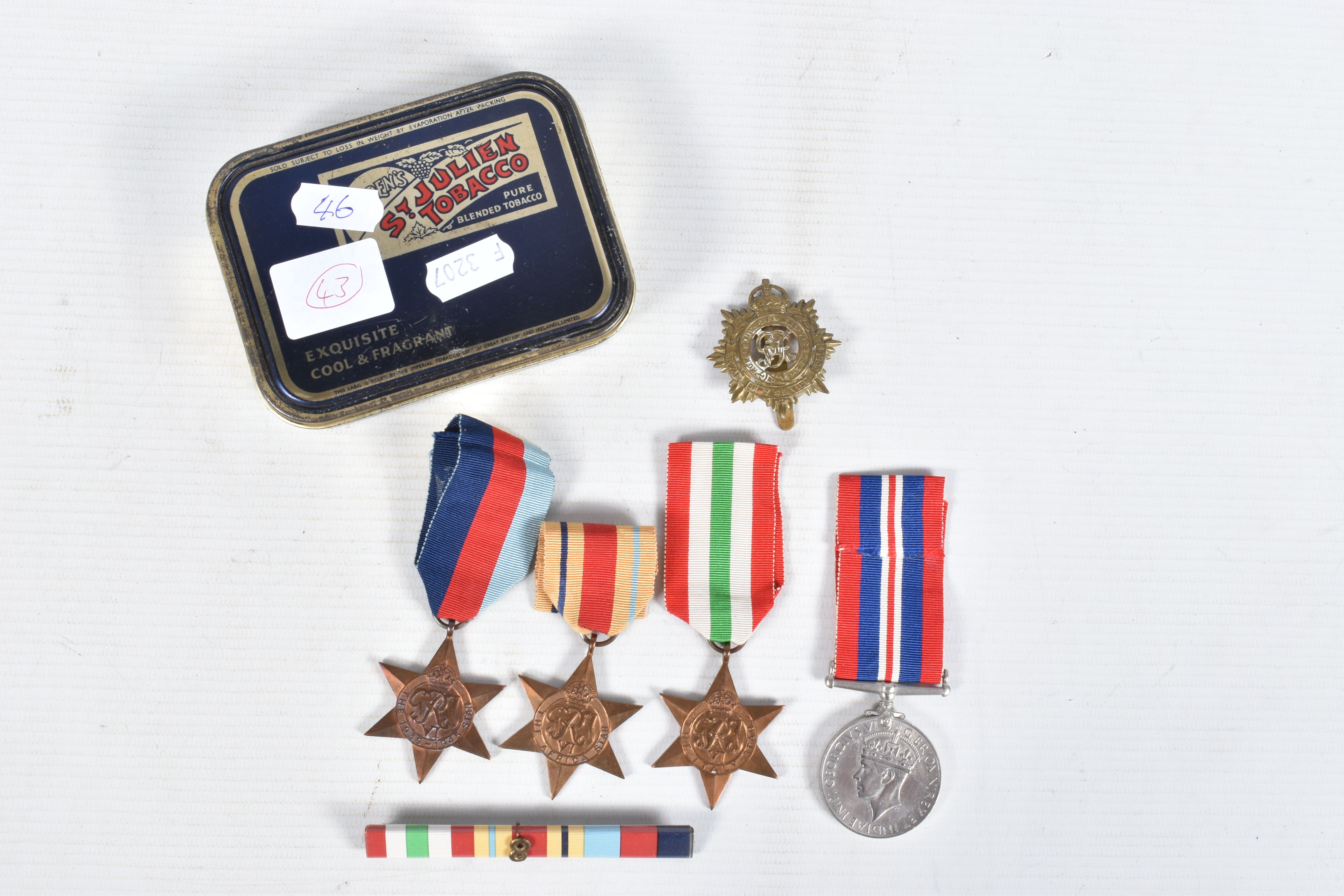 A TIN CONTAINING FOUR WWII MEDALS, a ribbon bar and a cap badge, the medals are all un-named and