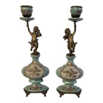 A PAIR OF BRONZE AND CERAMIC FIGURAL CANDLESTICKS The single sconces held aloft by cherubs
