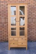 A light oak cabinet with twin glazed doors enclosing two shelves over four short drawers,