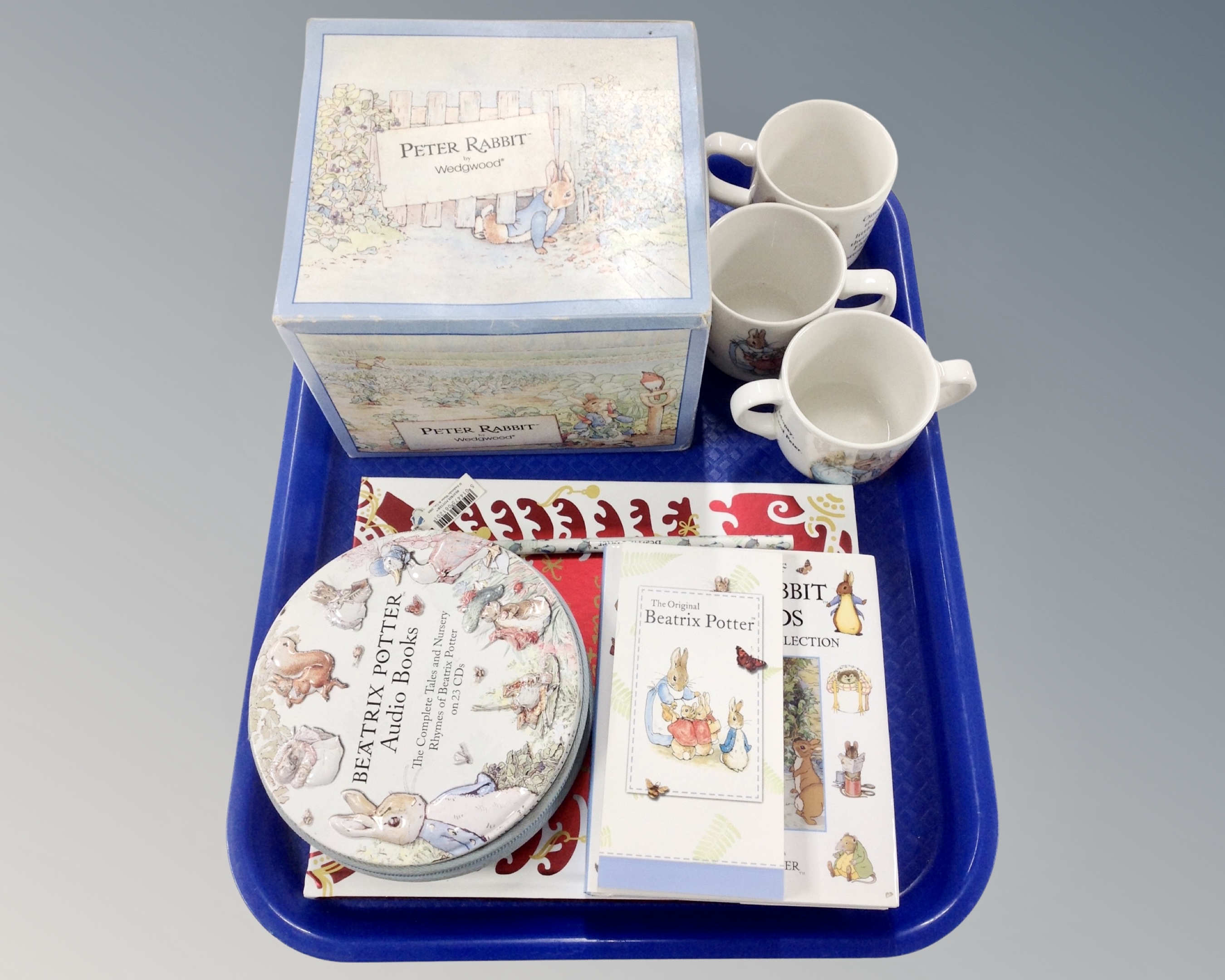 A tray of Wedgwood Peter Rabbit items including three mugs, boxed tea set, story books etc.