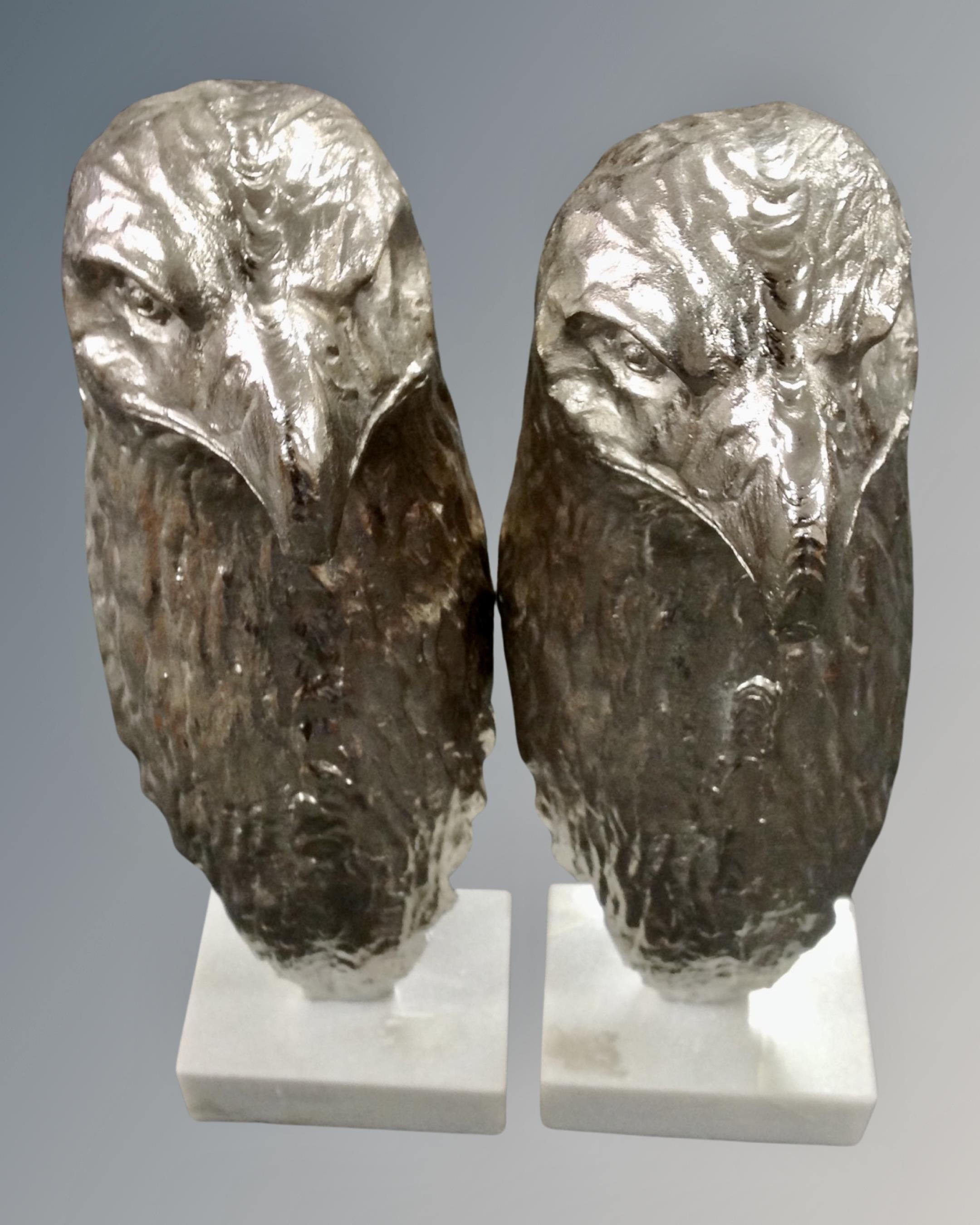 A pair of metal eagle head ornaments on white marble plinths.