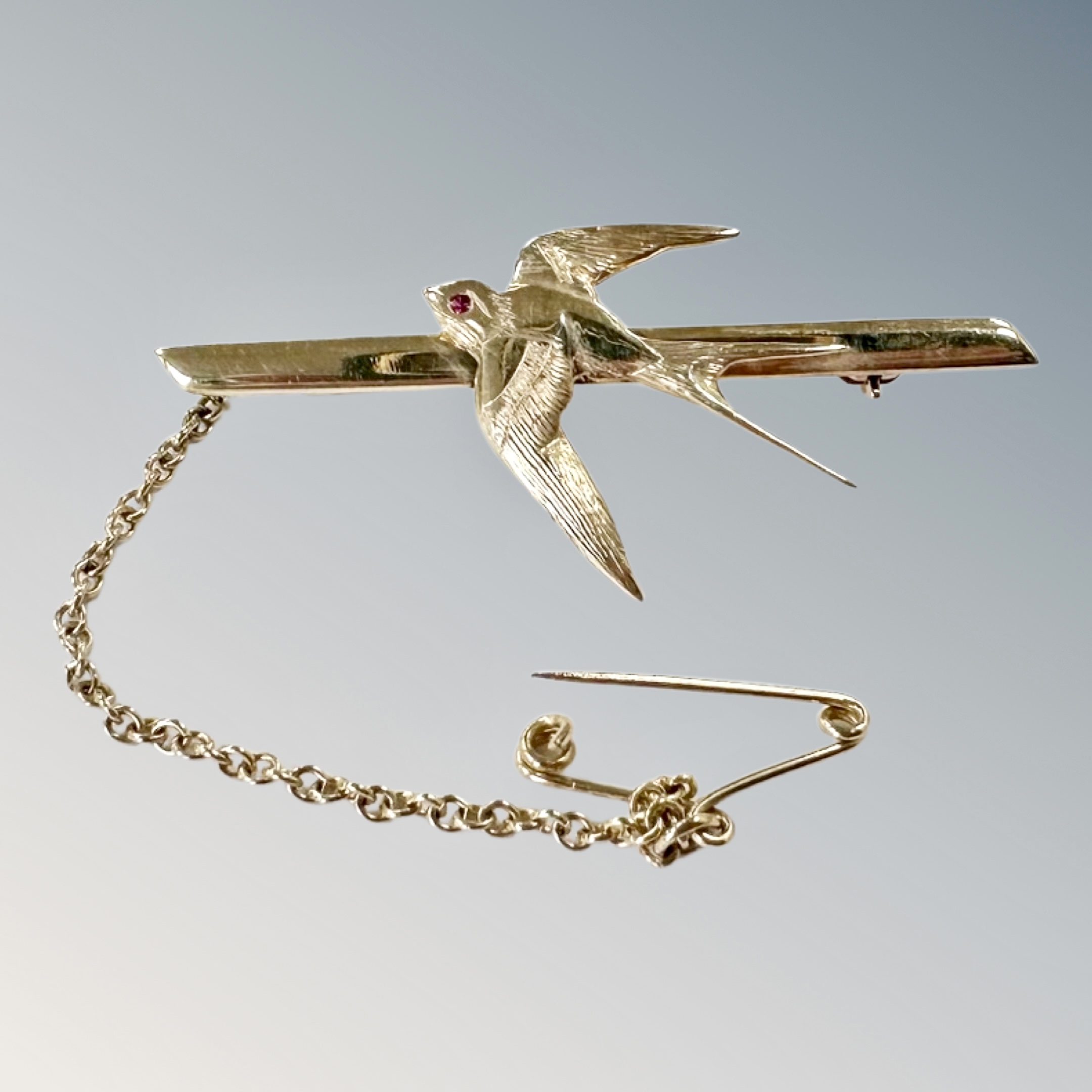 A 9ct gold swallow bar brooch with ruby-set eye, with safety chain, length 41mm.