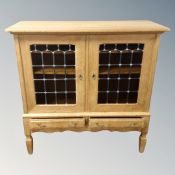 A blonde oak double leaded glass door side cabinet, fitted two drawers on raised legs,