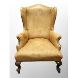 A George III style wingback armchair upholstered in a studded tan faux leather, raised on castors,